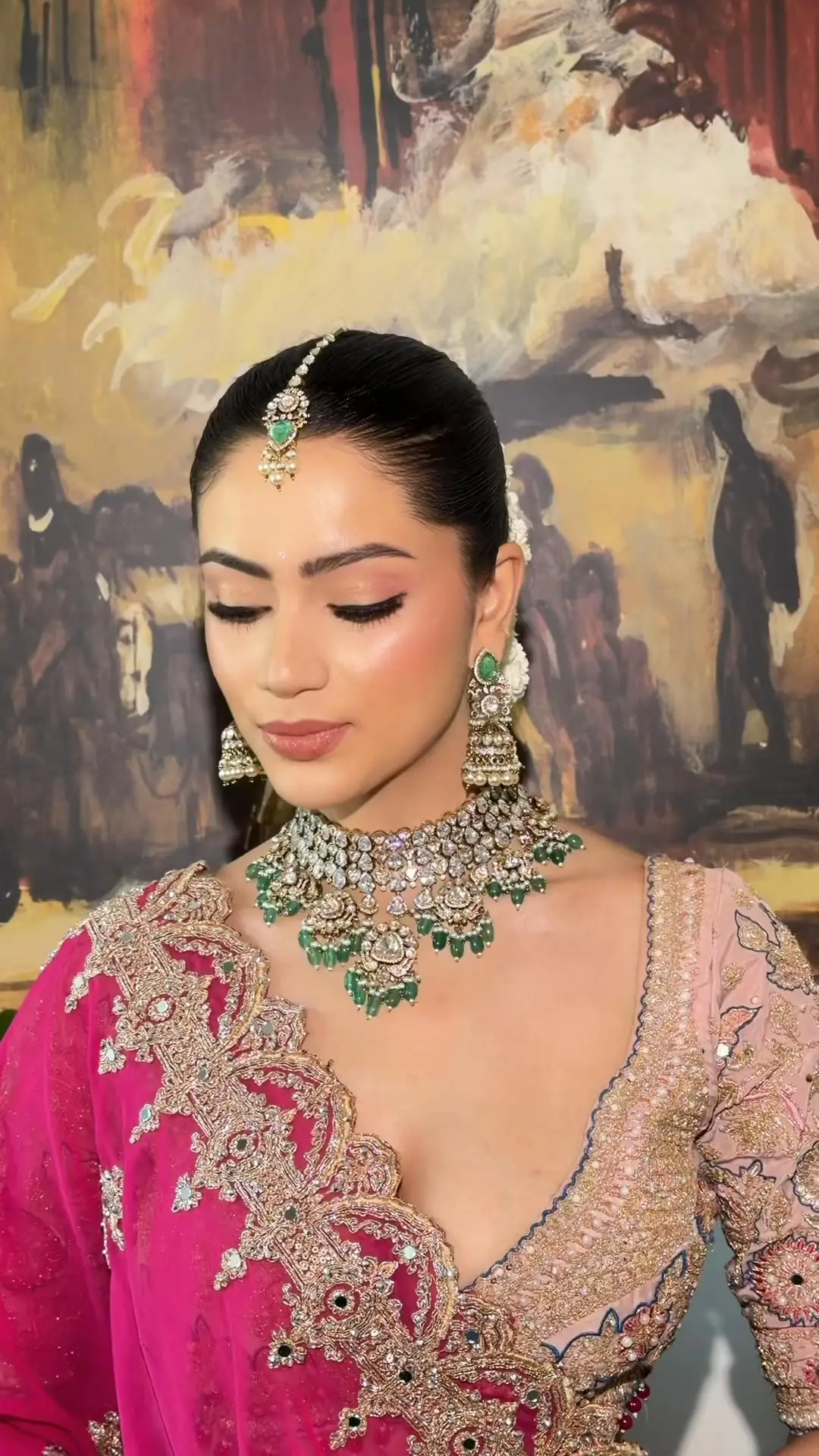 A bride looking down, eyes closed, wearing a diamond and emerald choker, jhumka earrings and a maang tikka, a pink and gold embroidered blouse, in front of a large painted mural.