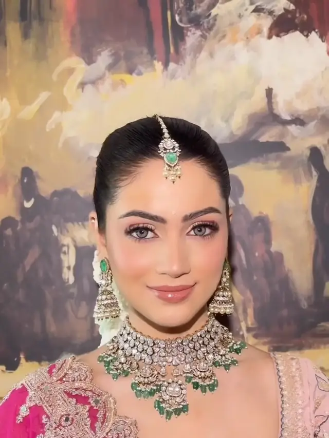 A bride smiling gently, wearing a gold and emerald maang tikka, a matching choker and jhumka earrings, a pink and gold embroidered outfit, in front of a large painted mural.