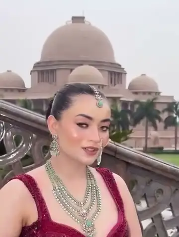 A bride in a maroon lehenga with a layered emerald and pearl necklace and a maang tikka, one hand raised to her hair, a domed palace building visible behind her.